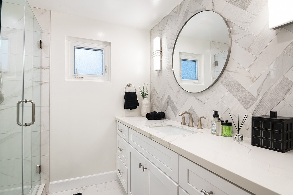 A bathroom with a white vanity, marble-look counter, round mirror and glass shower