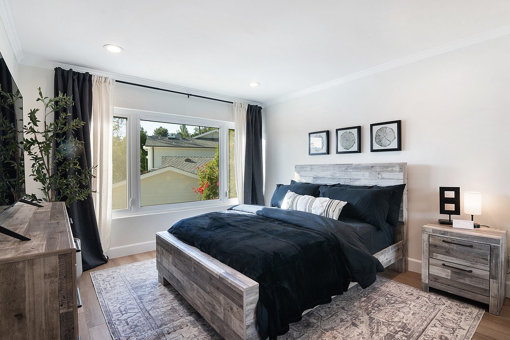 A bedroom with a queen bed, dark bedding and a wide curtained window