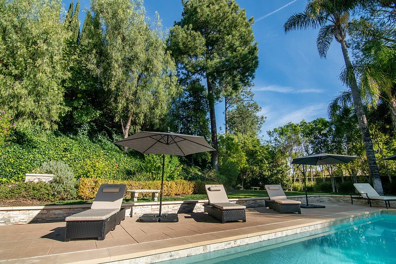 Cushioned lounge chairs and umbrellas beside the pool, backed by trees and garden