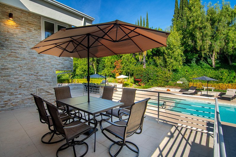 A shaded patio dining table beside the pool, with the garden beyond